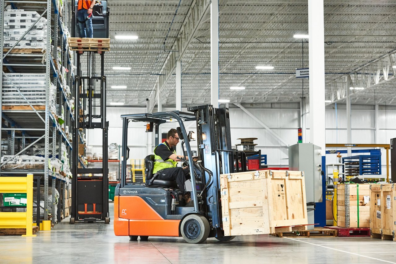 Man driving Toyota forklift in warehouse while carrying wooden crate 